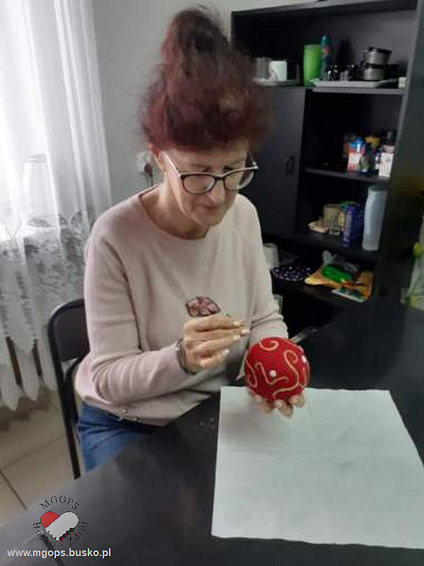 Seniorka dekoruje styropianową bombkę używając czerwonych i złotych dekoracji